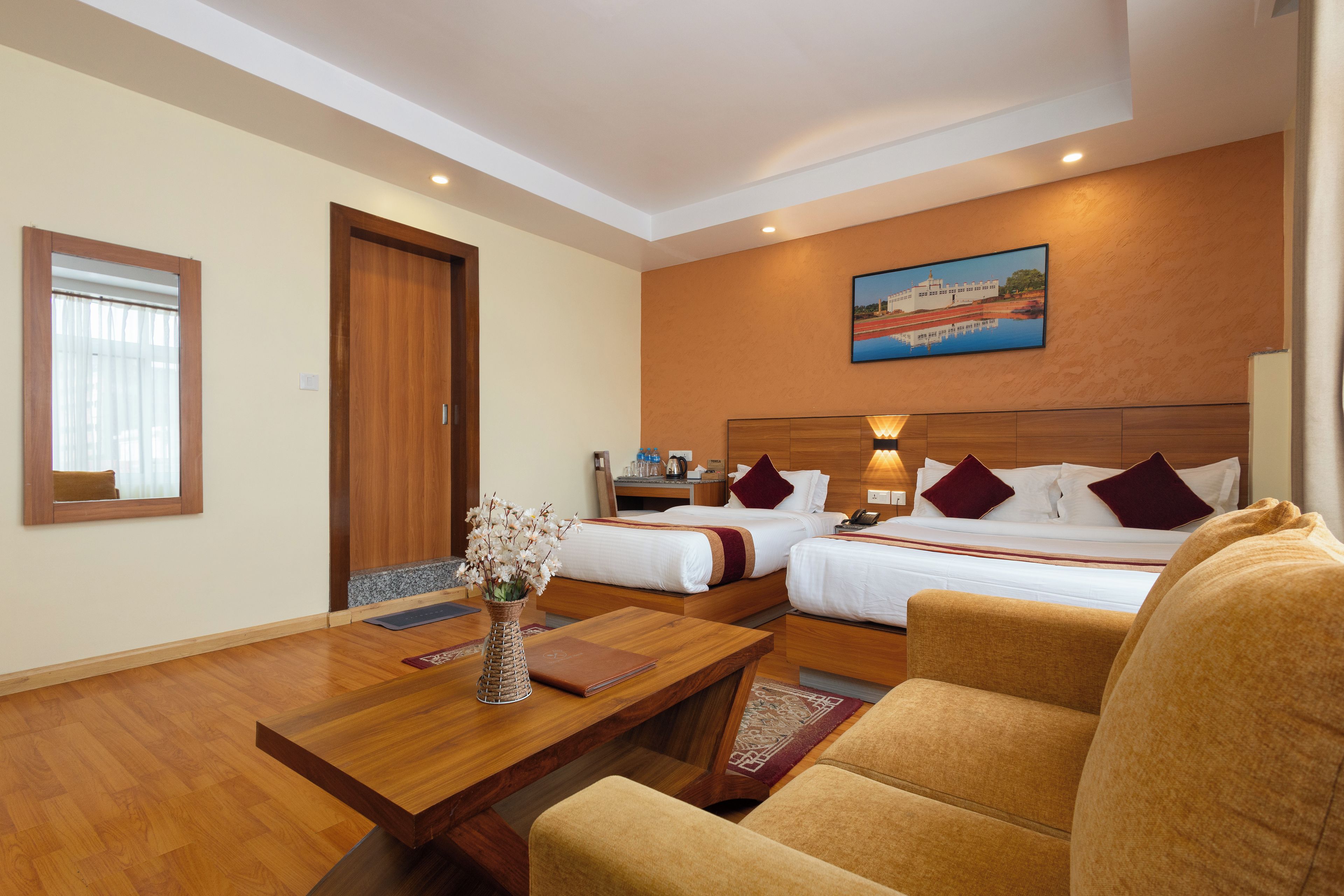 A hotel room with two beds, a wooden desk, a chair, a vase with flowers, and a painting on the wall.