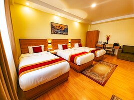 Habitación de hotel con tres camas, cabeceros de madera, un cuadro en la pared, un escritorio con silla y una mesita con un jarrón de flores.