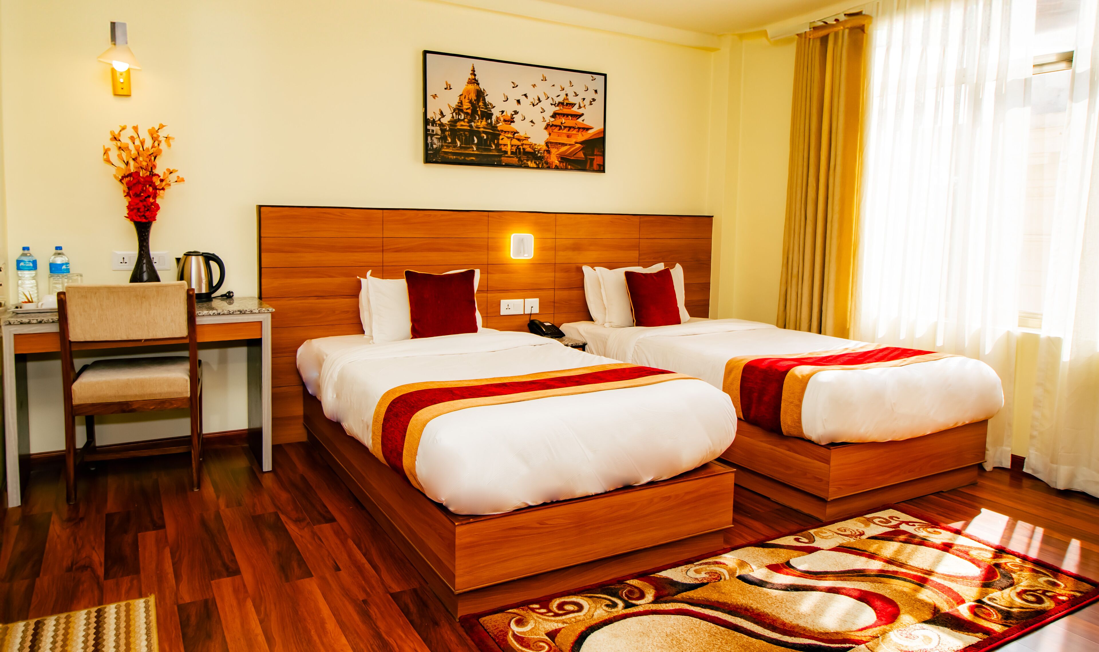 A hotel room with two beds, a desk, a chair, a vase with flowers, a painting of a temple, and a patterned rug.