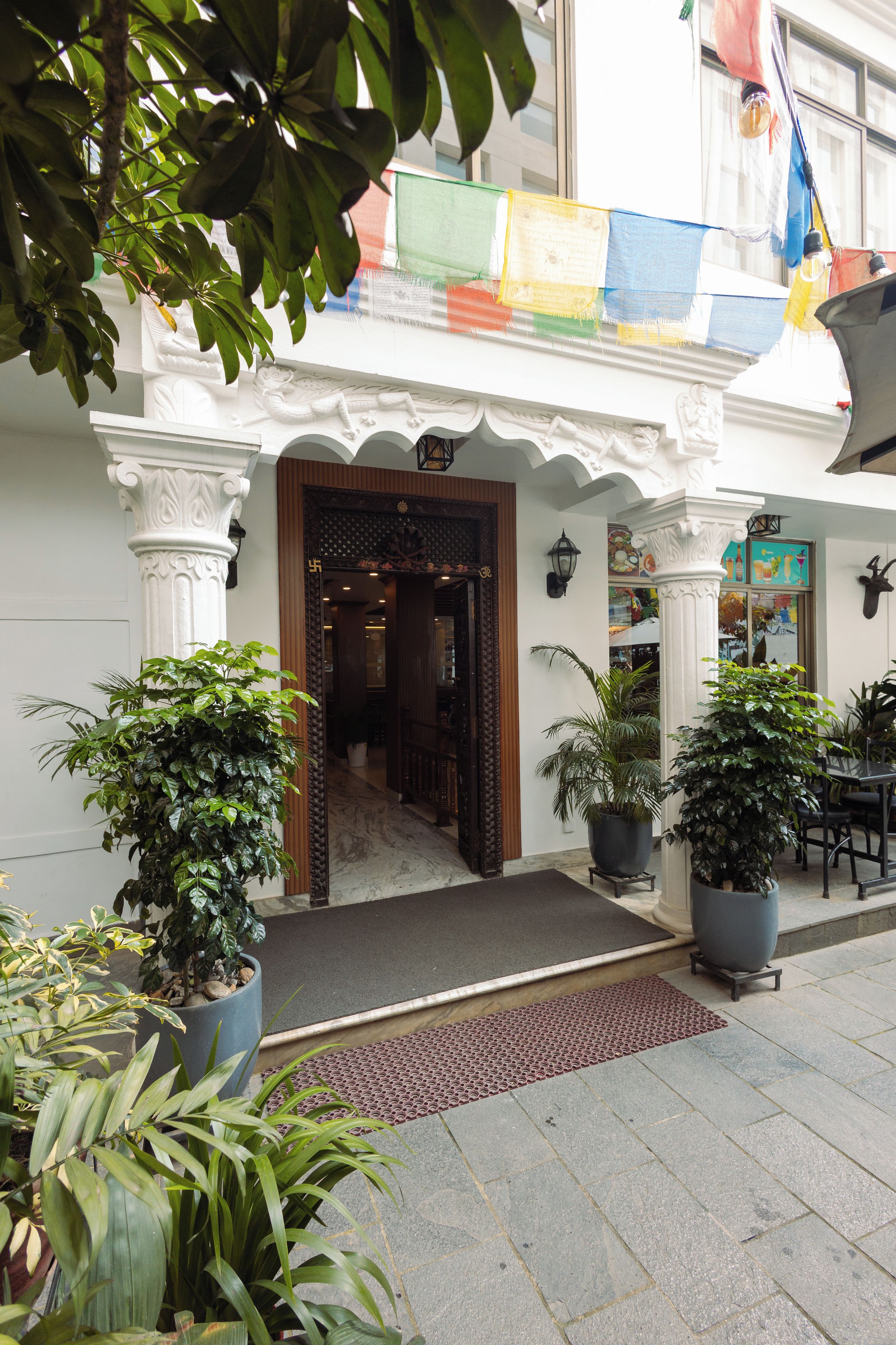 Una entrada de edificio con diseño tradicional, que incluye una puerta de madera y columnas blancas. Hay plantas en macetas frente a la entrada.