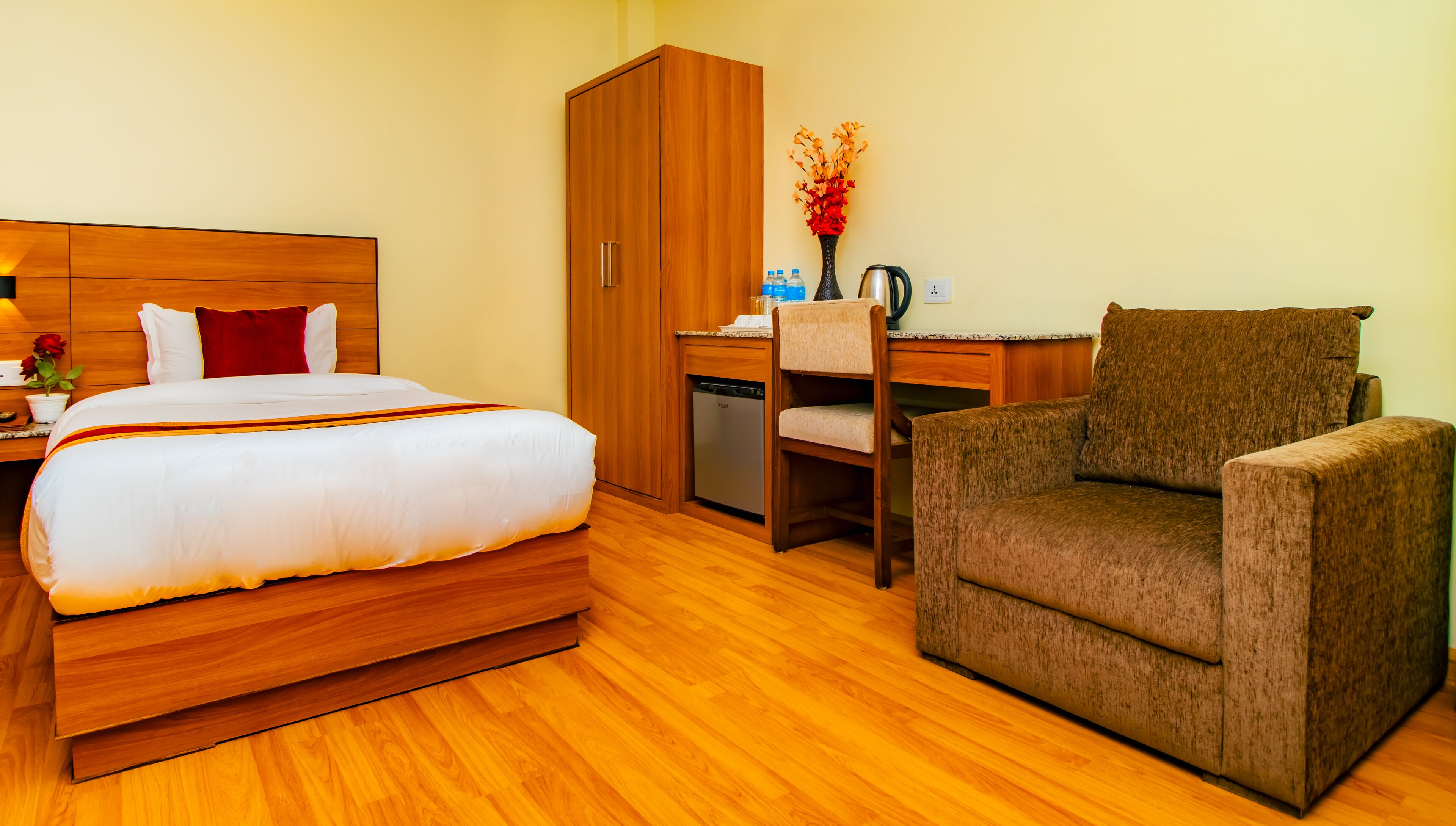 Une chambre d’hôtel avec un lit en bois, un fauteuil marron, un bureau avec une chaise et un petit meuble.