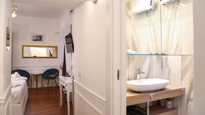 A hotel bathroom with a white sink, vanity, and a mirror. There is a chair and a small table with a television mounted on the wall. - Mama House Roma (Rome)