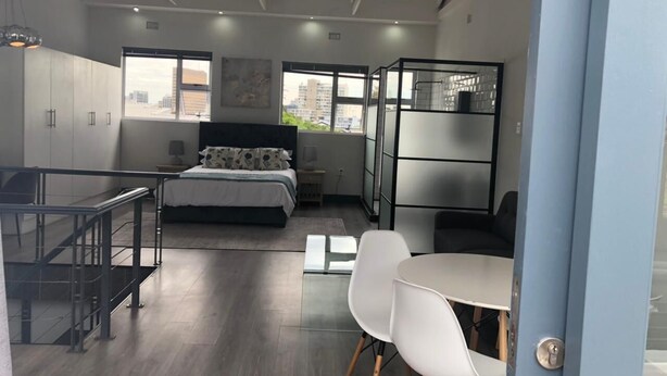 A modern bedroom with a bed, bedside table, and a view of the city through large windows. - Beautiful Newly Renovated Penthouse in the Trendy Gardens Surbub (Cape Town)
