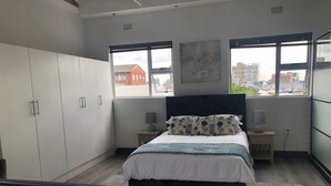 A modern bedroom with a large bed, two bedside tables with lamps, a wardrobe, and a view of buildings through large windows. - Beautiful Newly Renovated Penthouse in the Trendy Gardens Surbub (Cape Town)