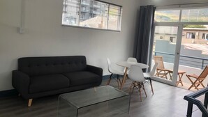 A modern living room with a dark sofa, a glass coffee table, and a dining area with white chairs and a small table. - Beautiful Newly Renovated Penthouse in the Trendy Gardens Surbub (Cape Town)