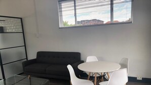 A modern living room with a dark grey sofa, a glass coffee table, and white chairs around a round table. - Beautiful Newly Renovated Penthouse in the Trendy Gardens Surbub (Cape Town)