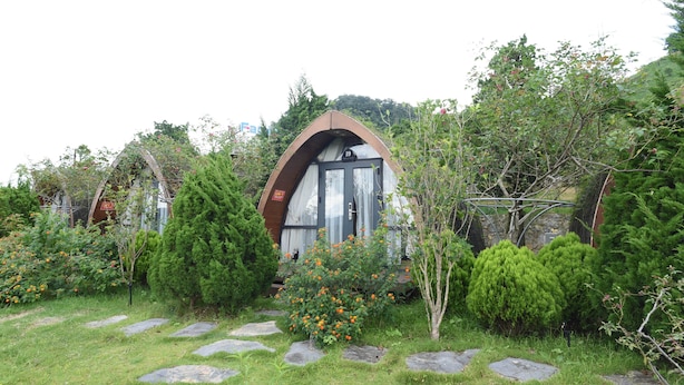A cozy cabin with a distinctive arched entrance, surrounded by lush greenery and a stone pathway. - Family 64 Home (Van Ho)