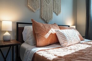 Een opgemaakt bed met een houten hoofdbord, twee bedlampjes en macramé wanddecoraties.