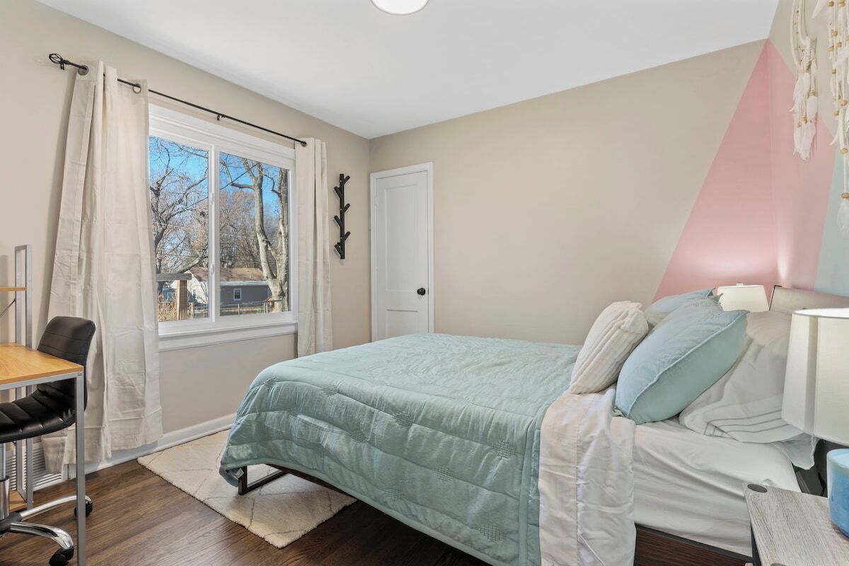 A bedroom with a bed, a desk, and a chair. There is a window with curtains, a door, and a wall-mounted coat rack.