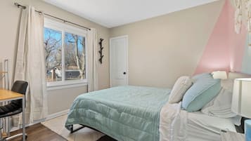 A bedroom with a bed, a desk, and a chair. There is a window with curtains, a door, and a wall-mounted coat rack.