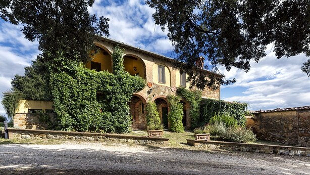 Un edificio storico con finestre ad arco e facciata in pietra, immerso nel verde e con un vialetto sterrato.