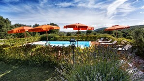 Area piscina con ombrelloni arancioni, poltrone relax e vista libera su vegetazione e colline circostanti.