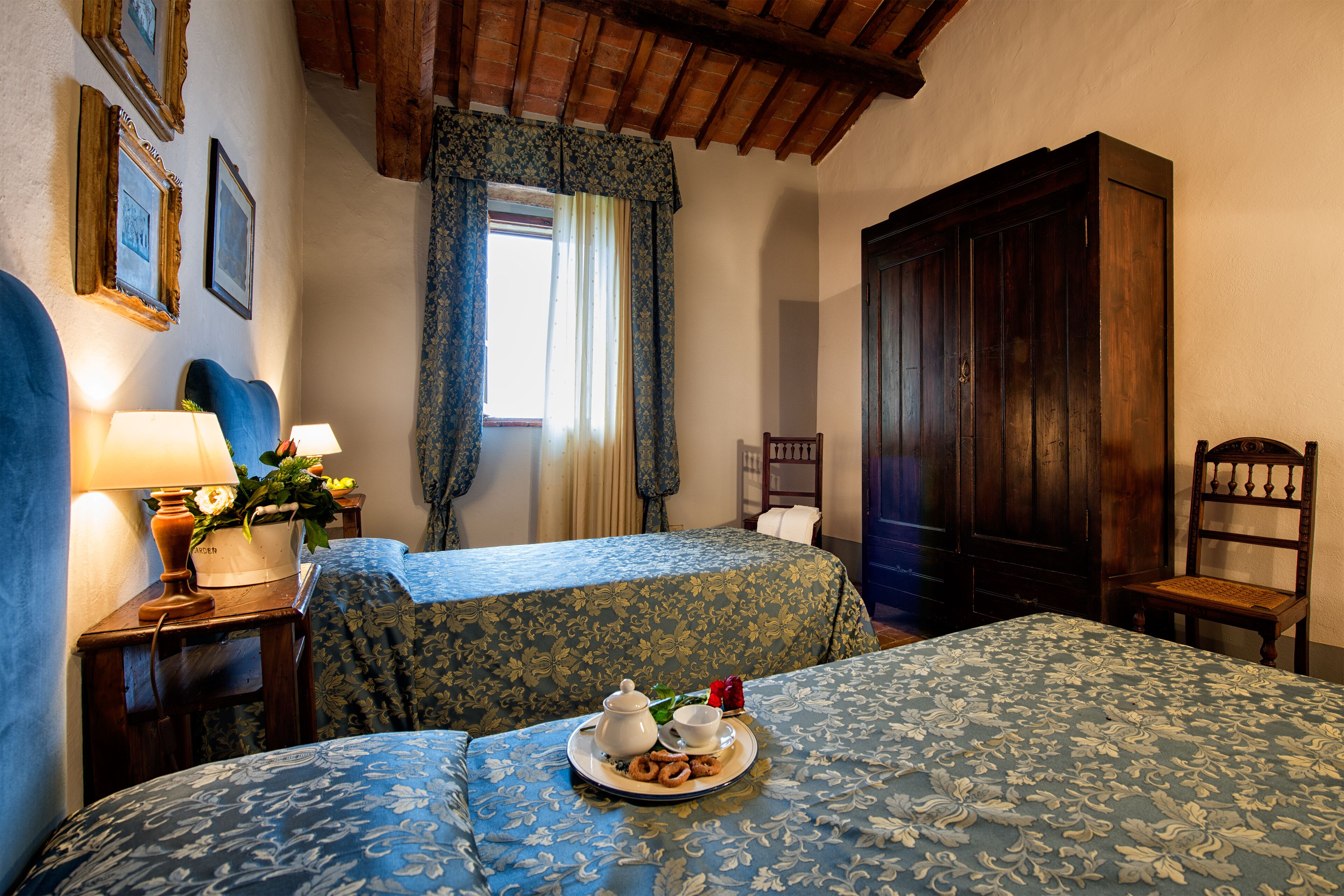 A hotel room with two beds, a wooden wardrobe, a nightstand with a lamp, and a tray with breakfast items.