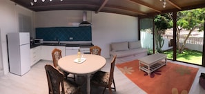Cozinha compacta com geladeira branca, micro-ondas e fogão. Há uma área de jantar com mesa redonda e cadeiras, e um sofá. O quarto possui uma janela grande que oferece vista para um jardim com árvores e uma cerca.
