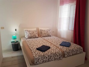 Un letto matrimoniale con biancheria a motivi, una lampada da comodino, un cestino e una finestra con tende rosse.