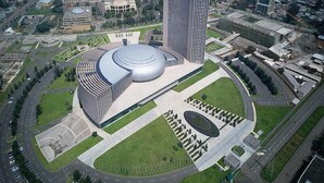 A modern architectural complex with a central circular building, surrounded by a landscaped area with a fountain and a parking lot.