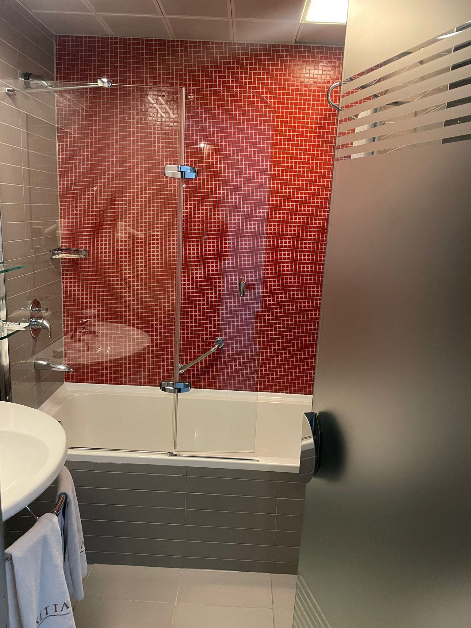 A modern bathroom with a red-tiled shower wall, a white bathtub, and a glass-enclosed shower.