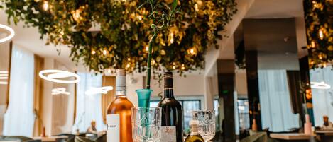 Una mesa con varias botellas y copas, probablemente para una cata de vinos, en un restaurante moderno con plantas colgantes e iluminación colgante.