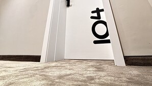 Un acceso estrecho y con ángulo, con una puerta blanca, un pomo negro y un símbolo con las letras 't' y 'o' en color negro en la puerta.