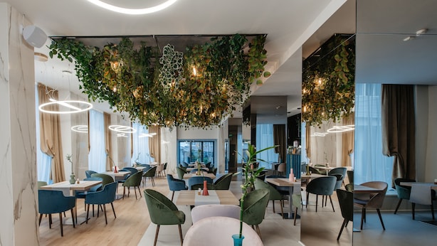 Un restaurante moderno con mesas de madera y sillas verdes, plantas colgantes e iluminación circular en el techo.
