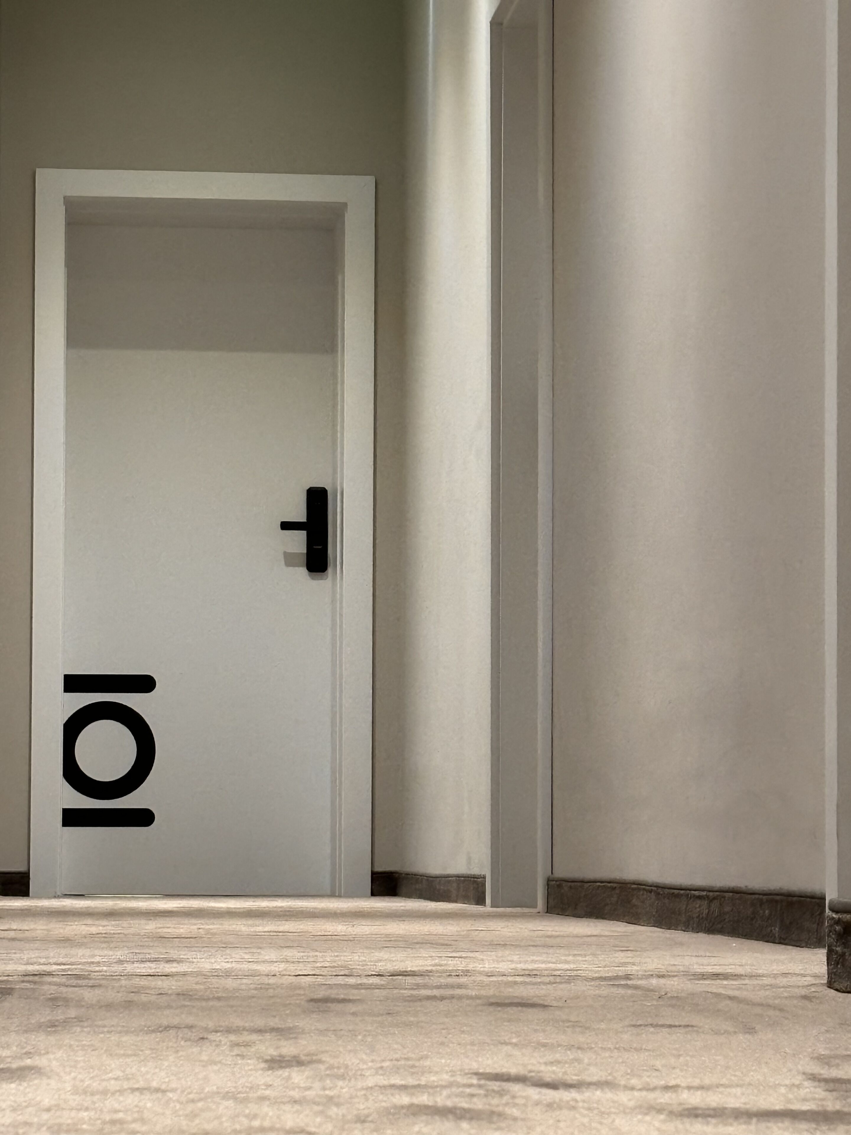 A hallway with a white door marked with a large '101' sign, a concrete floor, and a plain wall.