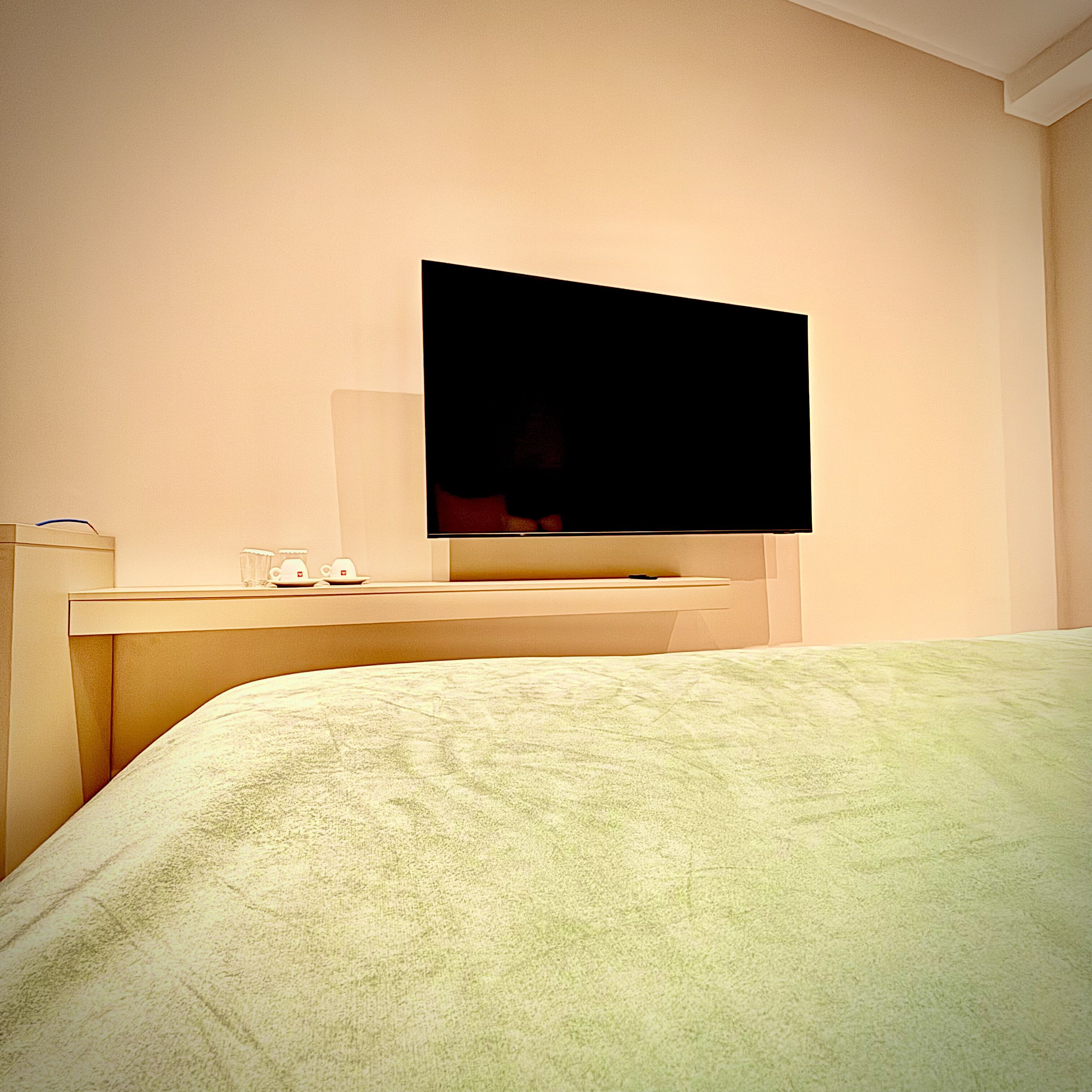A hotel room with a bed, a flat-screen TV mounted on the wall, and a bedside table with a cup and saucer.