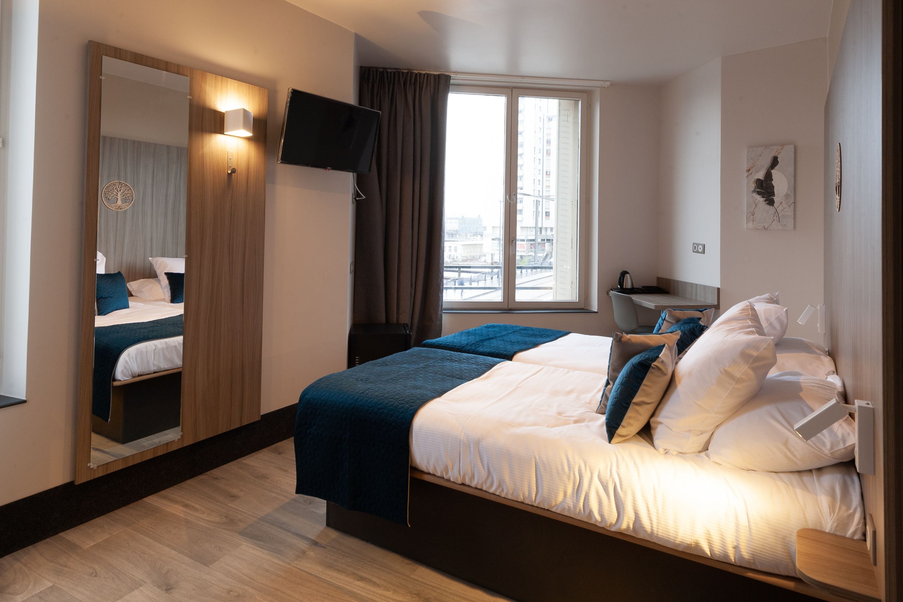 Habitación de hotel con cama, televisión, ventana con cortinas, una mesita pequeña y una obra de arte colgada en la pared.
