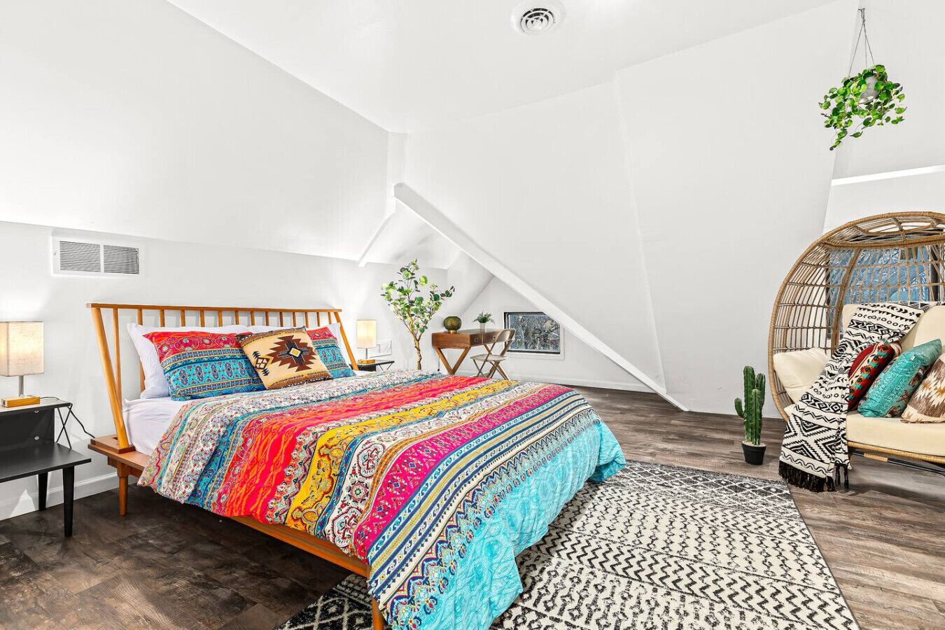 A bedroom with a wooden bed, colorful bedding, a hanging plant, a wicker chair, and a cactus.