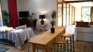 Een gezellige woonkamer met een houten eettafel, een bank en een televisie op een houten kast. Er staan potplanten en een lamp op een bijzettafeltje.