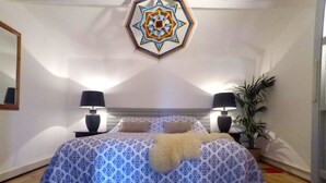 Een slaapkamer met een bed met een patroon, twee bedlampen, een decoratieve wandhanger en een plant in een pot.