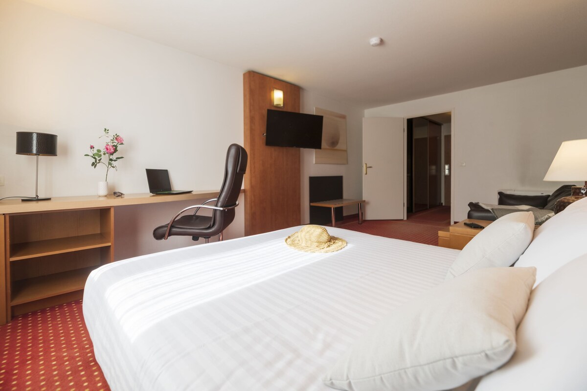 A hotel room with a large bed, a desk with a laptop, a chair, a TV mounted on the wall, and a bedside table with a lamp.