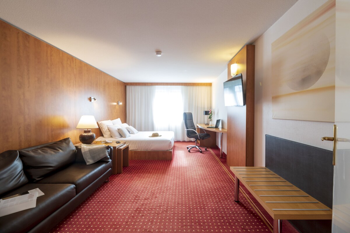 A hotel room with a wooden wall, a red carpet, a bed with white linens, a black leather sofa, a desk with a lamp, and a TV on the wall.