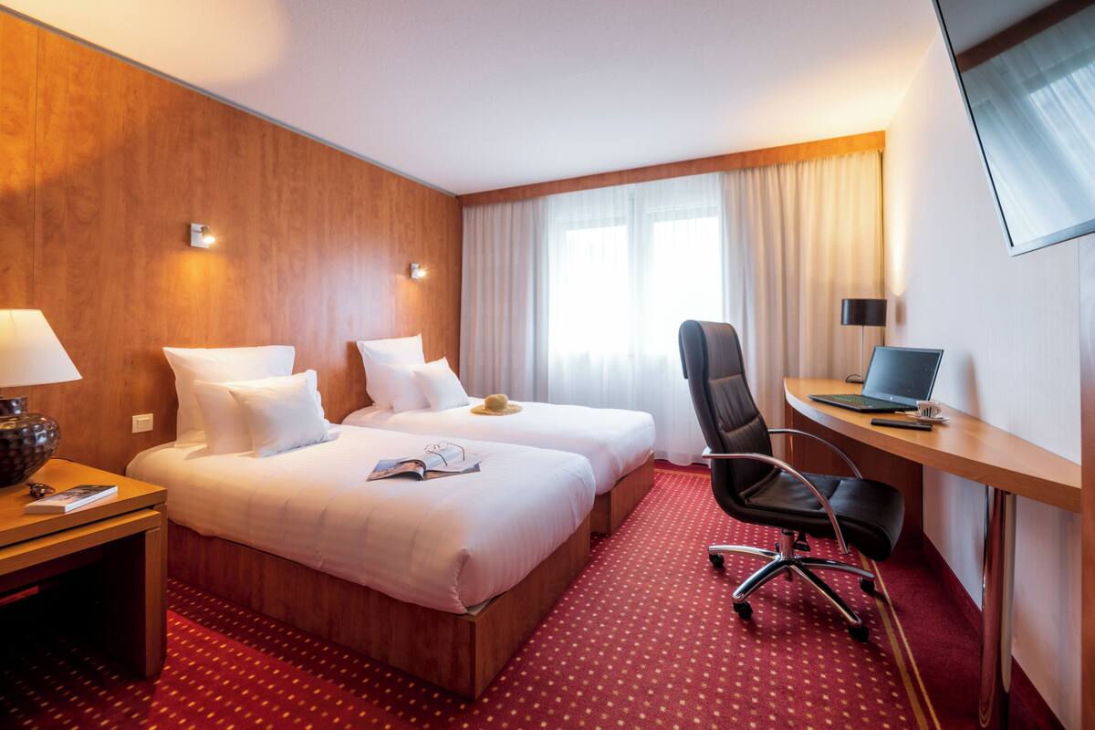 Habitación de hotel con dos camas, un escritorio con una computadora portátil, una silla y una mesita de noche con una lámpara.