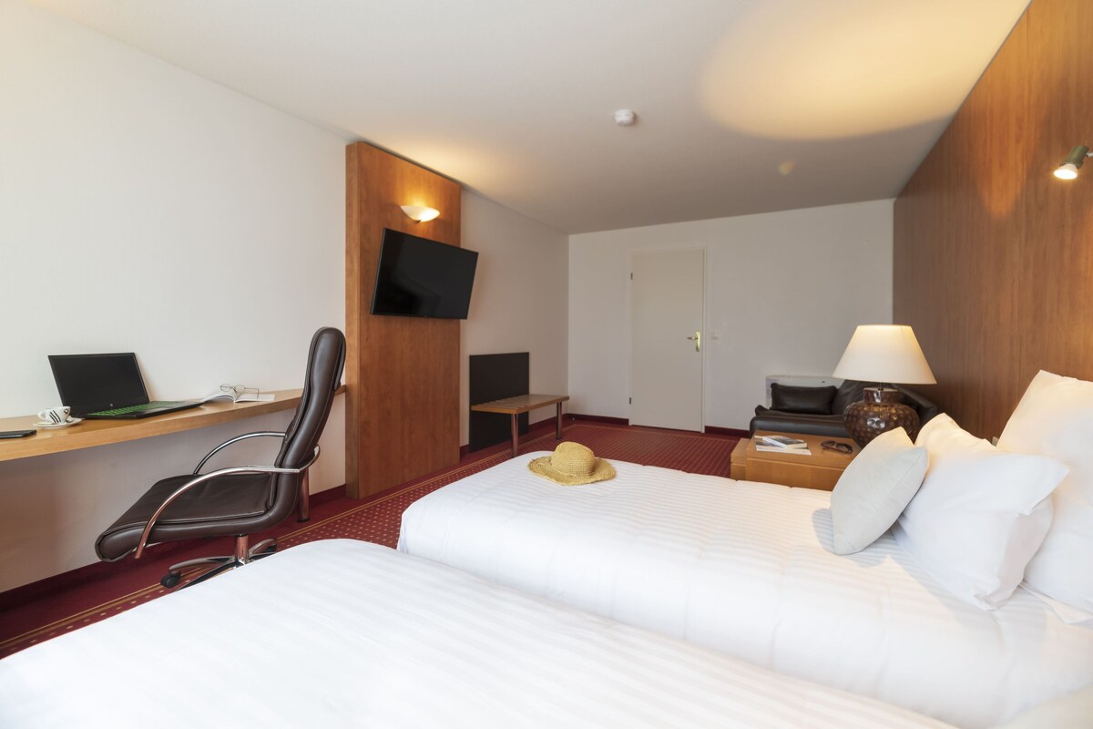 Ett hotellrum med två sängar, ett skrivbord med en bärbar dator, en stol, en TV, ett nattduksbord med en lampa och en dörr.