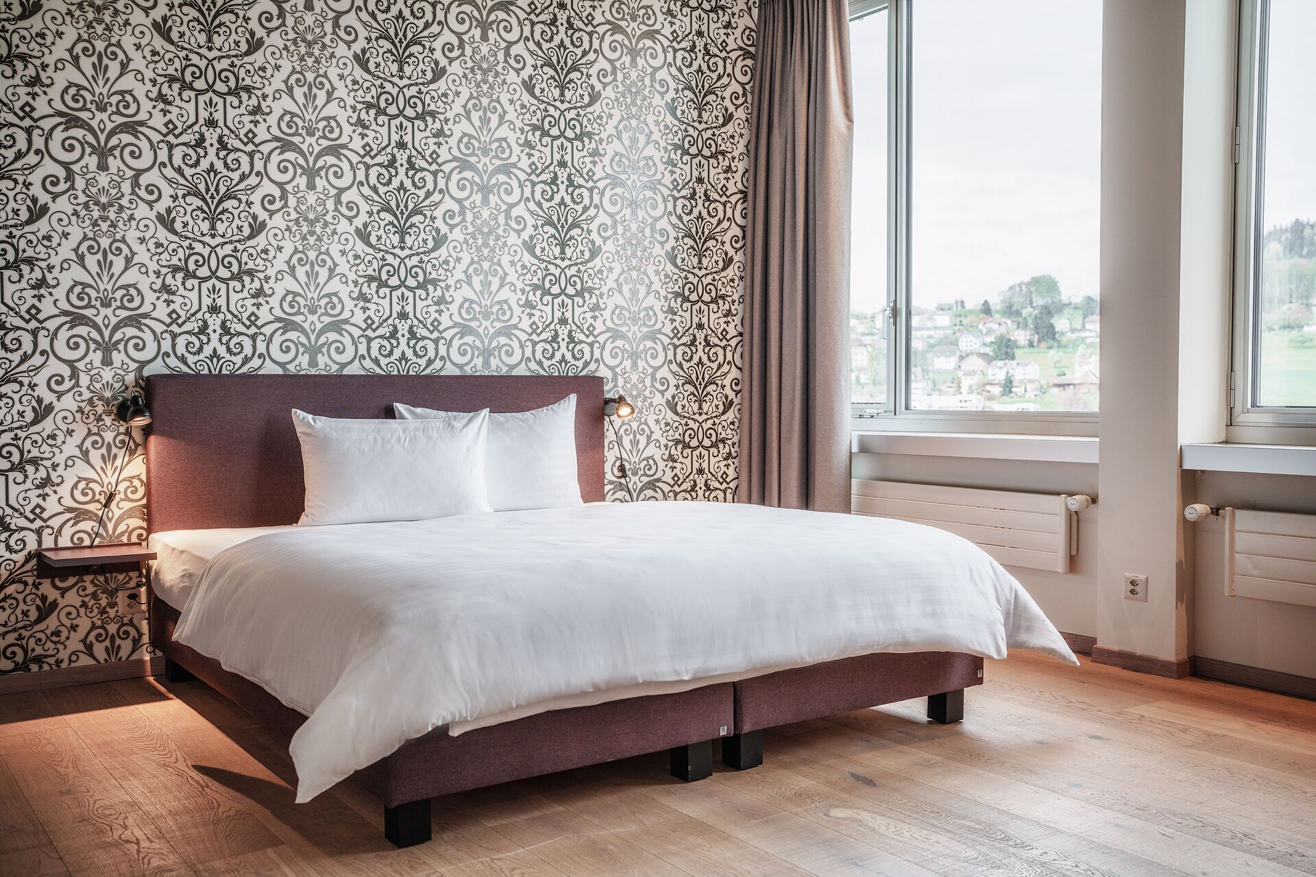 Ein ordentlich bezogenes Bett in einem Zimmer mit gemustertem Wandpapier, einem Fenster mit Vorhängen und einer Heizung.