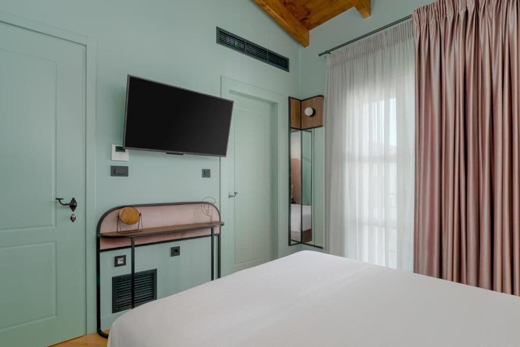 A bedroom with a bed, a television mounted on the wall, a mirror, and a window with curtains.