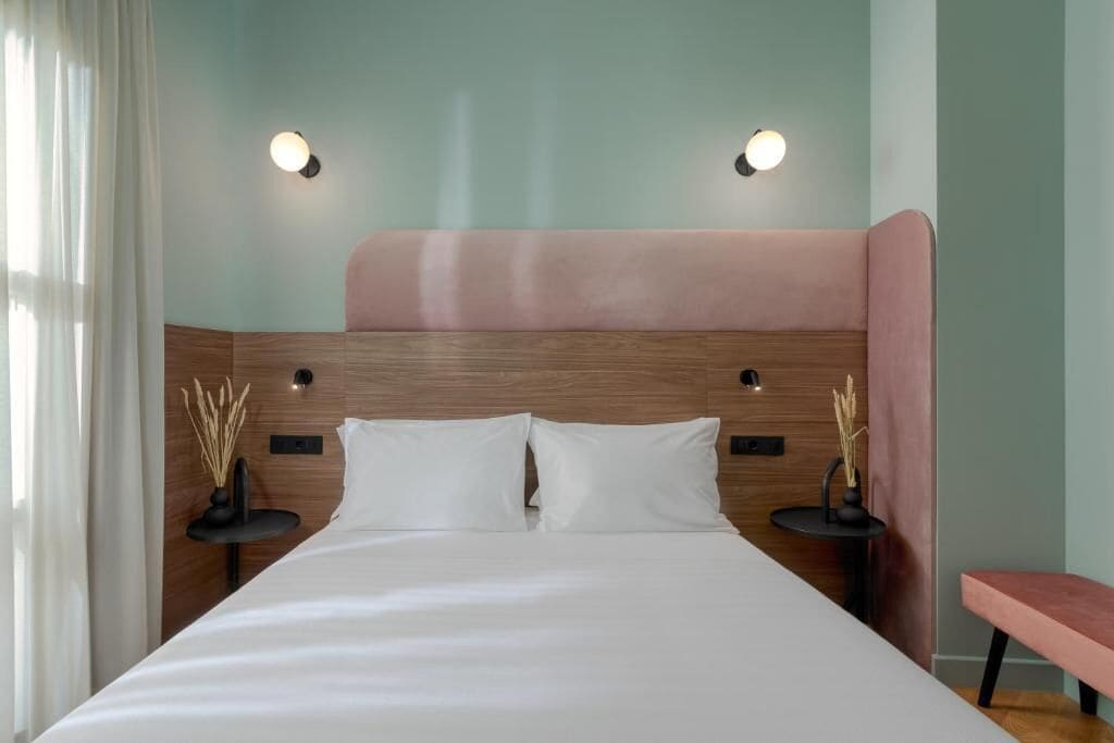 A hotel room with a large bed, two bedside tables, a bench, and wall-mounted lights.