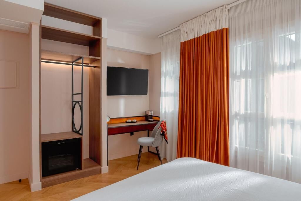 A hotel room with a bed, a desk with a chair, a TV mounted on the wall, and a wardrobe.