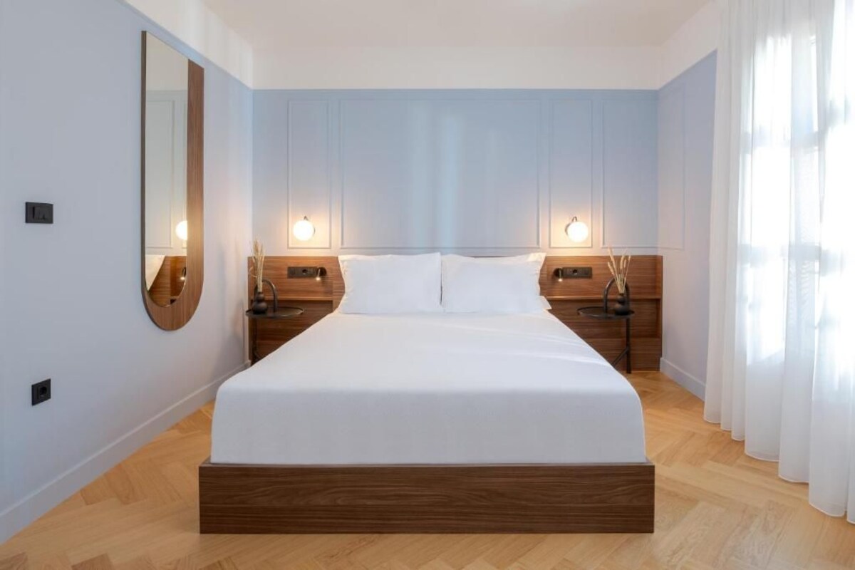 A modern hotel room with a large bed, wooden headboard, two bedside tables with lamps, and a large mirror on the left wall.