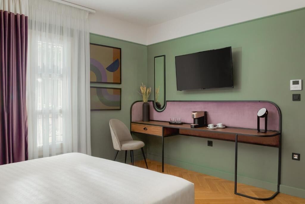 Ett hotellrum med en säng, ett skrivbord med en stol, en platt-TV och konstverk på väggen.