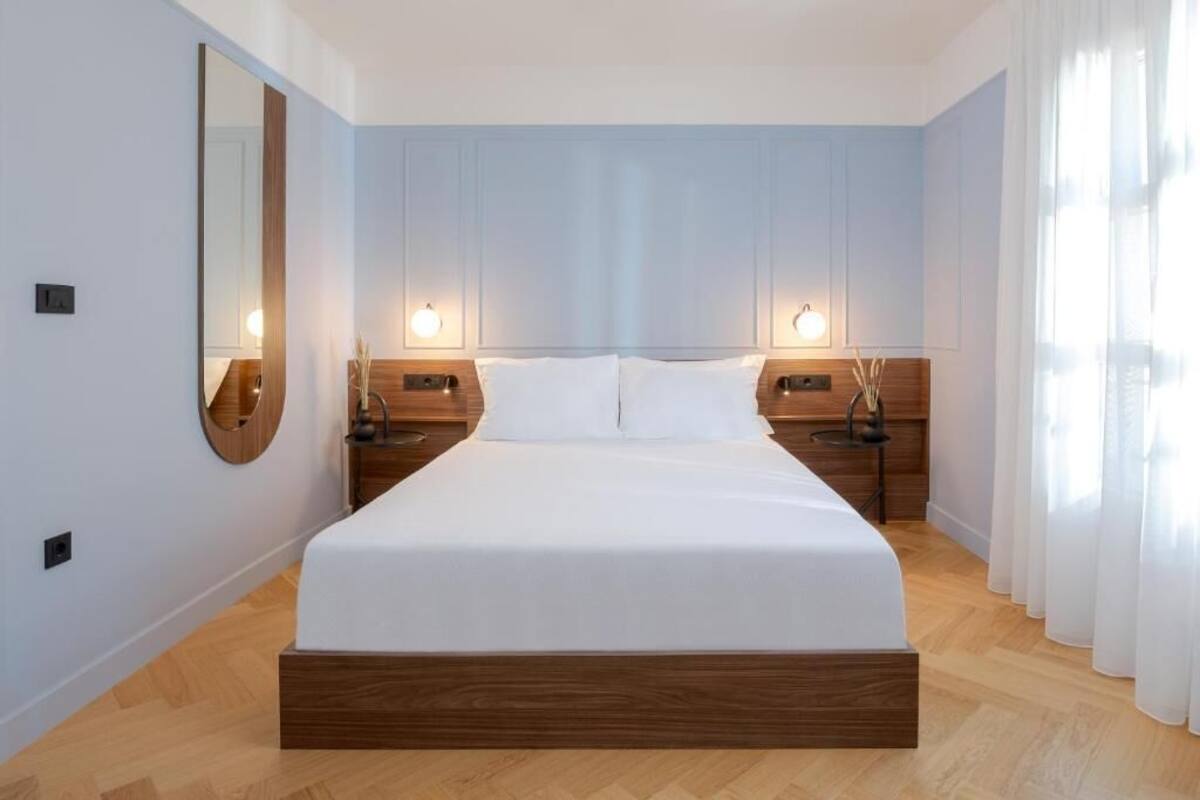 Een moderne hotelkamer met een groot bed, een houten hoofdeinde, twee nachtkastjes met lampen en een grote spiegel aan de linker muur.
