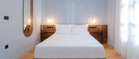 Quarto de hotel moderno com uma cama grande, cabeceira de madeira, duas mesas de cabeceira e um espelho amplo.