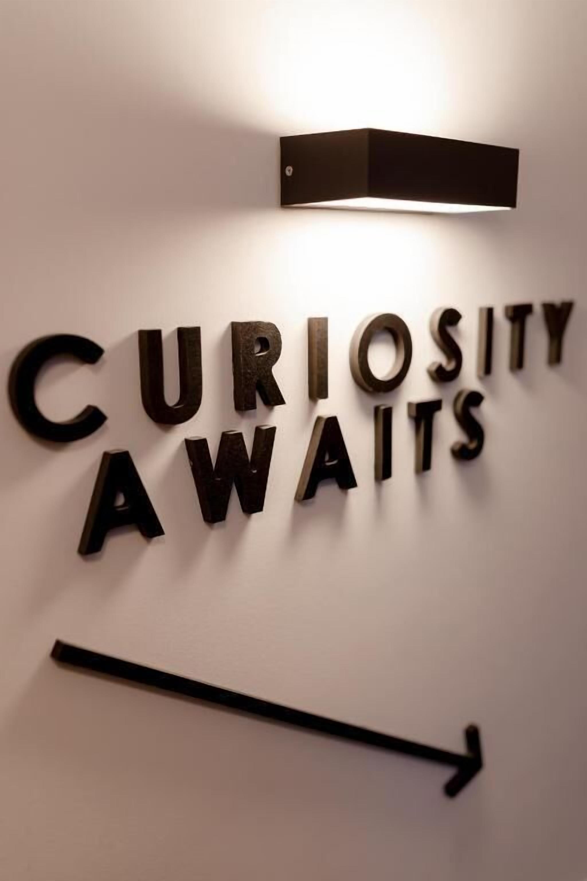 A wall with the words "CURIOSITY WAITS" illuminated by a light fixture.