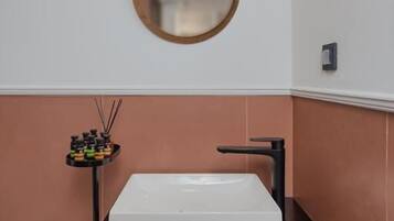 Un baño con un lavamanos blanco sobre un soporte de metal negro, un espejo redondo encima y una pequeña repisa con artículos de aseo.