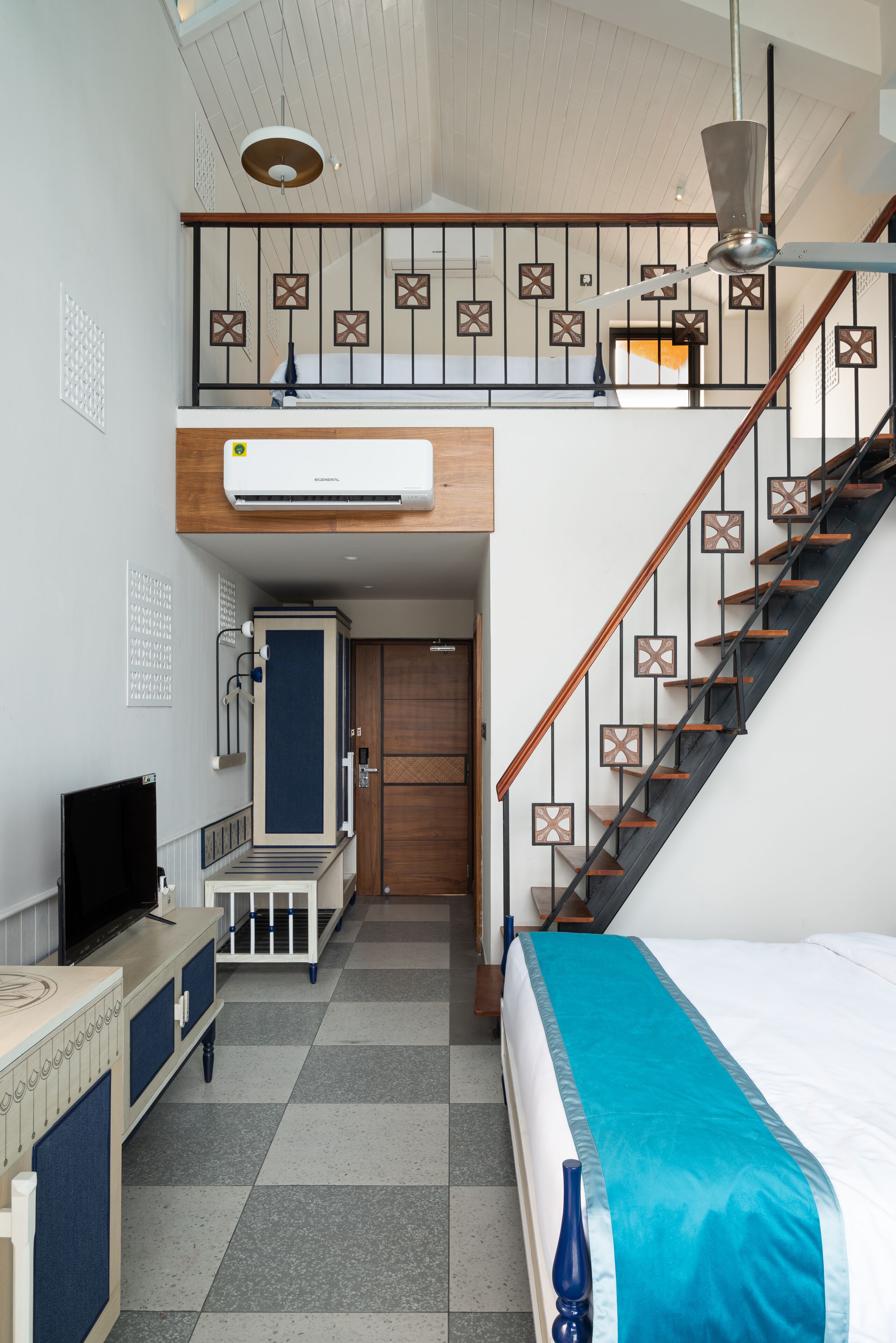 A compact hotel room with a lofted sleeping area, a staircase, and a built-in air conditioning unit.