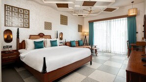 A hotel room with a large bed, wooden headboard, decorative wall panels, ceiling fans, a dining area with a table and chairs, and a large window with curtains. - Hotel Sonar Bangla Mayapur (Krishnanagar)
