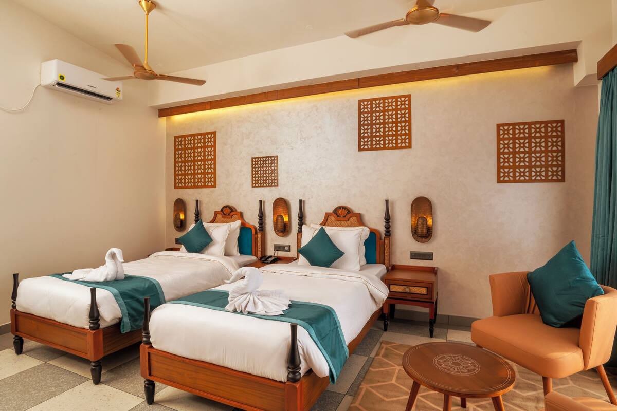 Quarto de hotel com duas camas, um espaço de estar, ventoinha no teto, unidade de ar condicionado e painéis decorativos na parede.