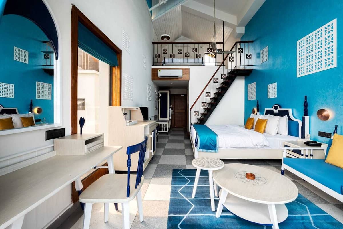 Quarto de hotel moderno com cama, escrivaninha, cadeira, mesa pequena e escada que leva a um andar superior.