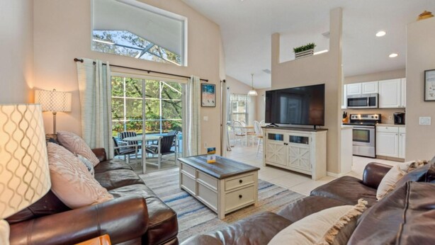 A spacious living area with a large leather sofa, a coffee table, and a TV. There is a kitchen area with white cabinets and a microwave.
