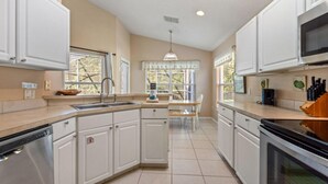A kitchen with white cabinets, a dishwasher, a microwave, and a stove. There is a window with blinds and a pendant light.